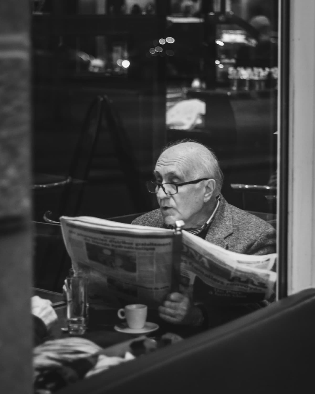 Older man in a blazer reading a newspaper at a cafe window